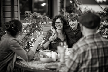 in the evening, dinner with the family around a table in the garden, in front of the wooden house. Everyone is having fun while a young boy gives his mother a hug.