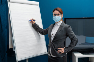 A woman in a medical mask leads a seminar in a conference hall. Business coach writes on a paper white board in the office. Female employee demonstrates presentation while maintaining social distance.