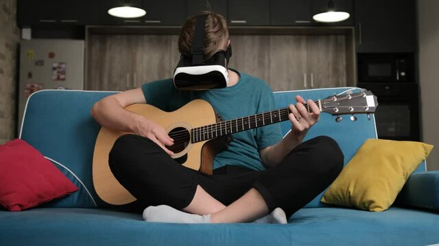 Man seated comfortably on sofa and wearing virtual reality glasses is playing at the guitar. He image how people see him