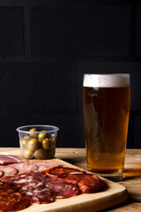 A cold glass of craft beer with a portion of snacks of cold cuts with olives, parma ham and salamis on wooden table over black background. Happy hour time.