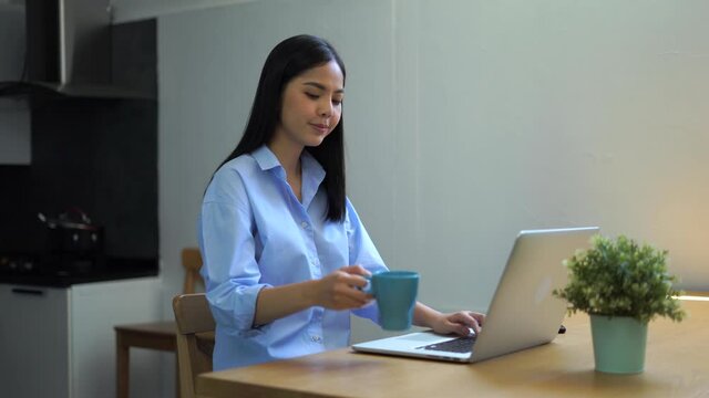 Work from home concept. Asian businesswoman working on laptop computer in the kitchen room and drinking tea or coffee. 4K resolution.