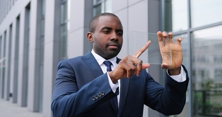African American young businessman in suit and tie standing at street and holding piece of glass on which tapping and scrolling like on screen. Touchscreen future technology. Transparent device.