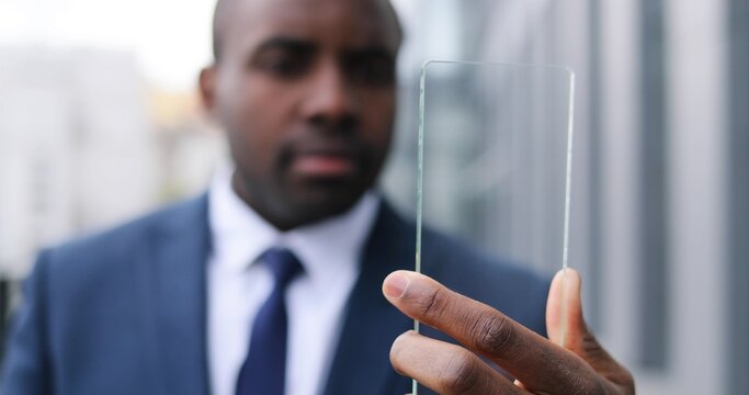 Close up of African American young businessman in suit and tie holding glass transparent screen, watching something. Phone screen. Outdoor. Future technology device. Futuristic technology. Blurred.