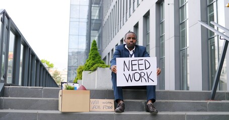 African American young sad and depressed man in despair sitting on stairs in city, showing poster Need work and Will work for food. Workless male looking anxious in depression Unemployment at lockdown