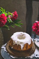 Bundt cake with white sugar glaze on wooden plate, rustic chair with peonies. Kitchen, natural light. Homemade dessert - tasty vanilla pie with icing. Copy space.