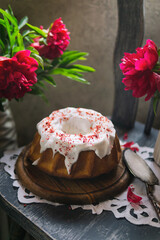 Bundt cake with white sugar glaze on wooden plate, rustic chair with peonies. Kitchen, natural light. Homemade dessert - tasty vanilla pie with icing. Copy space.