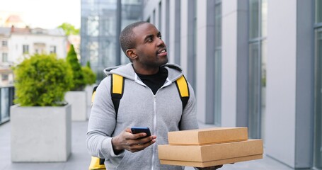 Fototapeta premium African American deliveryman with carton parcels standing the street, tapping on navigation map on phone looking for address. Male courier using navigating system on smartphone to deliver boxes.