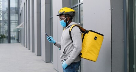 African American deliveryman in medical mask and casque standing outdoors at bicycle and texting message on smartphone. Bicyclist courier tapping, typing on mobile phone at street. Delivering concept