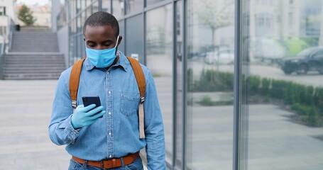 Obraz premium Close up of African American young man in medical mask walking the street and talking on mobile phone. Male pedestrian in respiratory protection strolling and speaking on cellphone. Conversation.