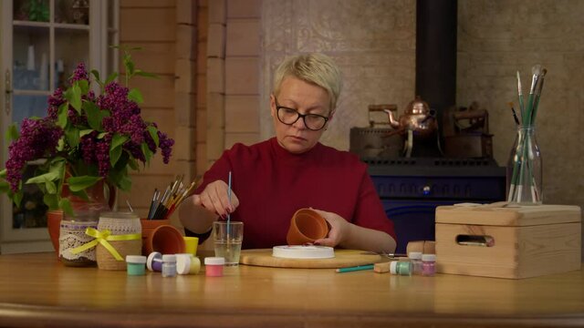 Caucasian focused middle aged female artist paints with paintbrush a clay pot in stylish art workshop. On the table are cans of oil paints and equipment for drawing. Wooden interior.