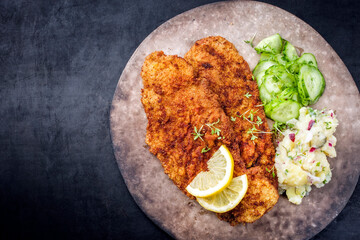 Traditional deep-fried schnitzel with potato and cucumber salad offered as top view on a rustic modern design plate with copy space left