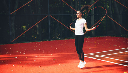 Fototapeta na wymiar Sporty caucasian girl using a jump rope on a red tennis stadium in a sunny summer morning
