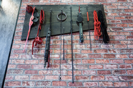A set of varied leather lashes for sexual pleasures hang on a wooden hanger on a brick wall. Sex equipment in red and black for role-playing games. BDSM room.