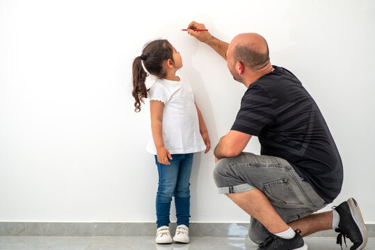 Father Measuring Height Of A Cute Child. Dad Measures The Growth Of Her Child Daughter At A Blank White Wall.