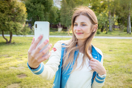 Young beautiful girl takes pictures of herself with a smartphone. Great summer weather. The woman is in the park, the street is sunny and warm. 