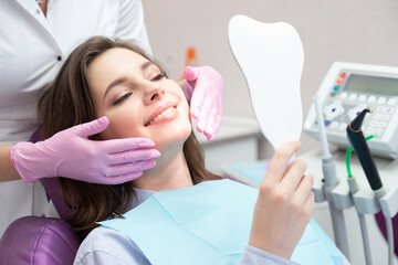 Young  smiling woman patient looking at mirror after teeth whitening procedure.