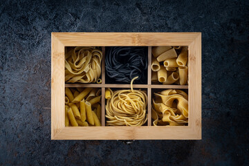 A wooden box with various types of dry pasta (spaghetti, rigatoni, tagliatelle, Penne, papardelle) on a dark stone table. Top view