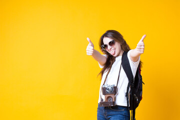 Cool travel tourist girl showing thumb up approval like gesture,satisfied recommend.Excited young woman in white t-shirt,backpack,sunglasses,retro photo camera isolated on yellow background.copy space