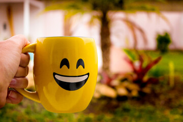 Mão segurando uma caneca de café com rosto sorrindo, lembrando um emoji. A cor da caneca é amarela e ao fundo há um jardim com palmeira e bromélias