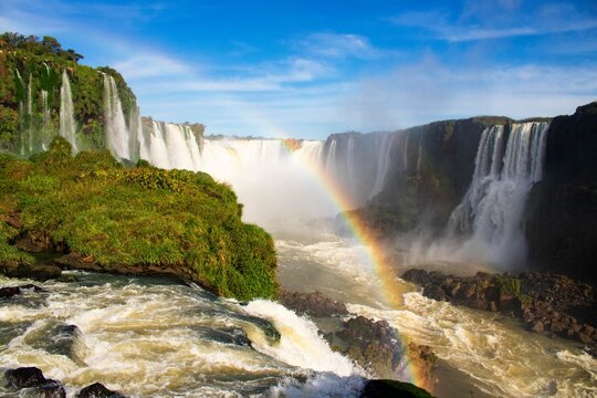 Cacheira, cataratas do Igua&ccedil;u. Em Foz do Igua&ccedil;u, Paran&aacute;, Brasil. C&eacute;u azul, dia ensolarado, um arco &iacute;ris &eacute; formado em frente as cataratas. Pode-se ver as rochas e natureza do local. Ponto tur&iacute;stico.