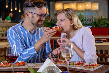 Happy young romantic couple in love feeding each other with bruschetta at dinner in a beautiful fancy restaurant.