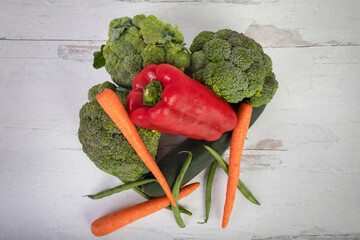 Overview of stacked vegetables green red and orange broccoli crowns red bell peppers and carrots on a white wood background flat lay