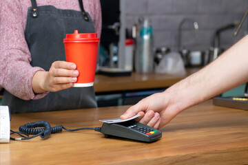 Takeaway coffee. barista holds out a red paper glass with a drink, the buyer pays with a card