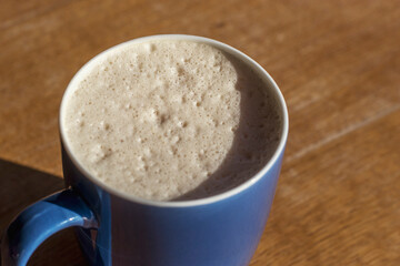 A glossy blue cup with latte coffee and airy foam stands on a brown wooden table. The sun shines brightly and objects on the table cast a clear shadow.