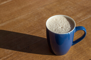 A glossy blue cup with latte coffee and airy foam stands on a brown wooden table. The sun shines brightly and objects on the table cast a clear shadow.