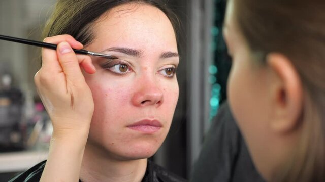 Professional makeup artist paints eyeshadows with brush to a young woman in a beauty salon. 4K slow motion footage