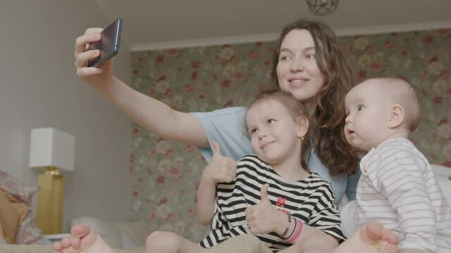 Young Mother and Two Daughters, Preschooler and Infant, Make Selfie with Smartphone, Lying on a Bed. Cozy Home Interior.
