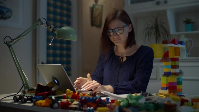 Young irritated 30s working from home mother with pile of kids toys on the working table with laptop and documents. Angry woman throwing away toys from working table. 