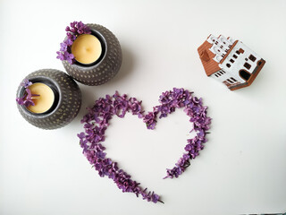 a heart of lilac flowers next to saffron candles and a white ceramic house. base for postcard