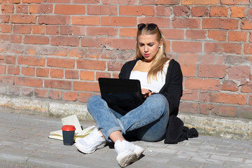 Fototapeta na wymiar A young girl with blond hair, dressed in jeans, a white T-shirt, black sweater and white sneakers, secluded herself against a brick wall for remote work during the period of self-isolation.