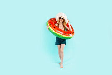 Full length, happy girl in sunglasses, swimsuit and summer hat, standing with an inflatable swimming circle, on a blue background