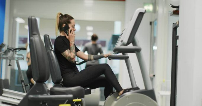 woman exercising with elliptical cross trainer while talking on cellphone at gym.Side view, slow motion. Woman training at elliptical bike while talking on mobile phone.Woman pedaling using smartphone