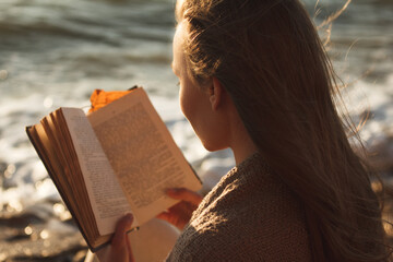 An attractive girl wrapped in a plaid sits on a stone by the sea at sunset and reads a book....