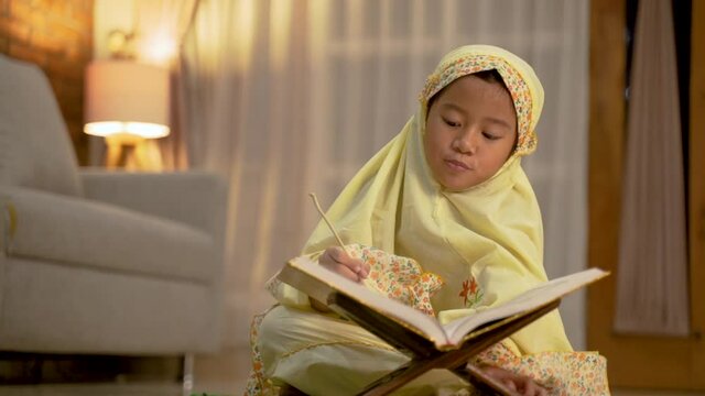cute asian girl reading quran by herself at home. muslim toddler with hijab