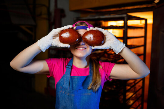 A professional baker woman holds fresh buns in her hands and hears their smell in the bakery. She is wearing a denim frock and a cap. Production of bakery products. Rack with hot crispy pastries.