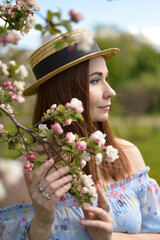 A beautiful girl in blue dress and straw hat holds flowering branch of Apple tree and looks into the distance in summer.