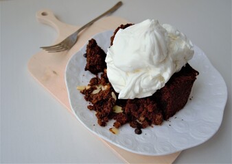 Homemade chocolate nut dessert with whipped cream on a white ceramic saucer and on a wooden board. In the background is a metal decorative fork (the focus is specially blurred)