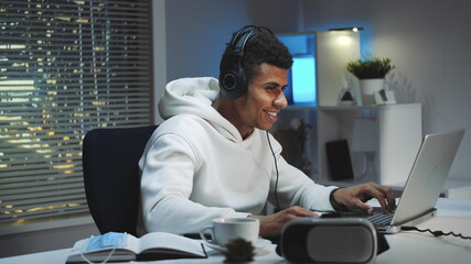 Close-up shot of Black gamer in white hoodie and with headphones playing games on computer in the...