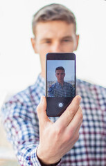 Caucasian  men  is making a selfie portrait with the help of smart phone. There is a city on the back ground.  Dressed into checkered shirt. Stands on the 9th floor.