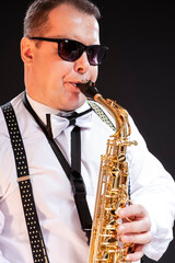 Fototapeta premium Music Concepts. Portrait of Passionate and Extravagant Caucasian Saxophonist in White Shirt Posing in Sunglasses Against Black Background.