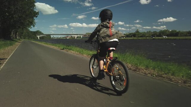 Little boy rides bike on paved path along lake, POV follow, 4k