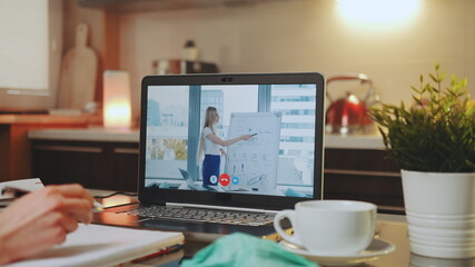 Online video conference on laptop with female speaker in home office. In the foreground: writing hands, medical mask and a cup.