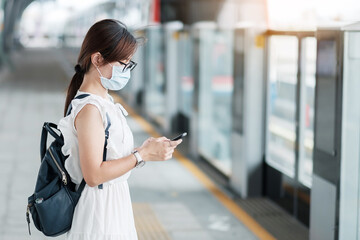 young Asian girl wearing Surgical face mask protect coronavirus inflection, Happy tourist woman using smartphone during walking in subway station. new normal and life after covid-19 pandemic - Powered by Adobe