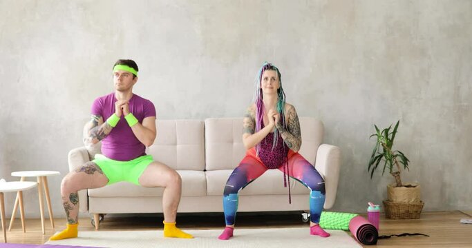 Ridiculous man and woman newcomers in sport in colourful sportswear making squats exercises at home, front view. Joke, mem, humor, parody, joking behavior. Fitness, workout, training together concept.