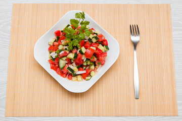 Fresh salad of finely chopped cucumbers, tomatoes and onions with sunflower oil in a white plate and a fork on a wooden table. The concept of healthy nutrition and eco-friendly, vegetarian diet.