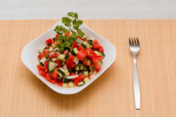 Fresh salad of finely chopped cucumbers, tomatoes and onions with sunflower oil in a white plate and a fork on a wooden table. The concept of healthy nutrition and eco-friendly, vegetarian diet.